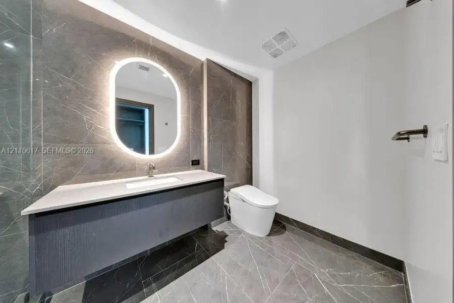 Floating vanity featuring a stone-topped counter and integrated sink, illuminated oval wall mirror, large-format stone tile wall and flooring, and wall-mounted toilet Floating vanity featuring a stone-topped counter and integrated sink, illuminated oval wall mirror, large-format stone tile wall and flooring, and wall-mounted toilet