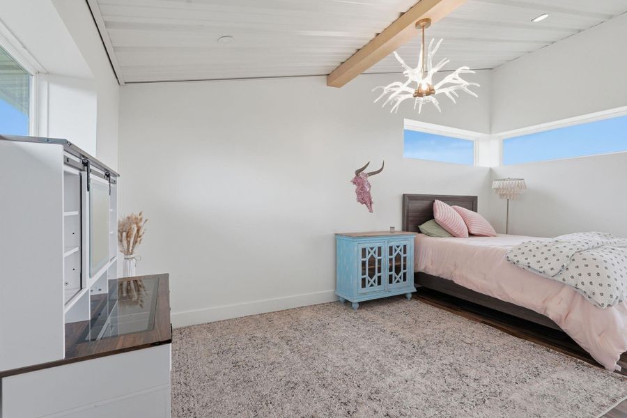 This bedroom features a modern, minimalist design with white walls and a carpeted floor. It includes a single bed with pink bedding, a unique antler-style ceiling light, and a blue bedside cabinet. Natural light streams through high, horizontal windows, creating a bright and airy atmosphere.