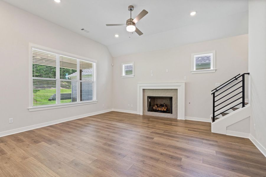 Spacious living area featuring wood-finish flooring, a fireplace with a white mantel, and a staircase with a black metal railing Spacious living area featuring wood-finish flooring, a fireplace with a white mantel, and a staircase with a black metal railing