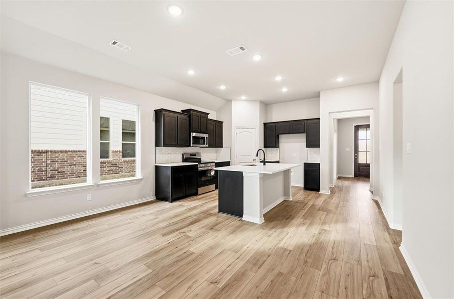 Kitchen featuring appliances with stainless steel finishes, a center island with sink, light wood-style floors, dark cabinetry, and decorative backsplash