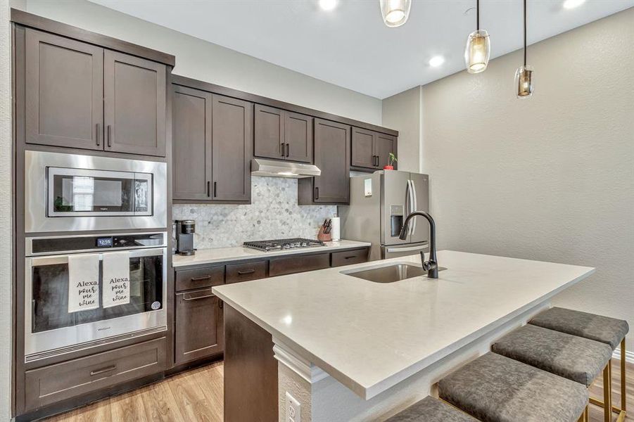 Kitchen featuring light hardwood / wood-style floors, tasteful backsplash, hanging light fixtures, a kitchen island with sink, and appliances with stainless steel finishes