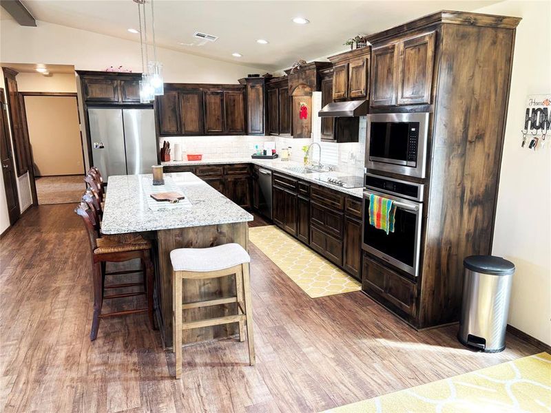 Kitchen with tasteful backsplash, light stone countertops, vaulted ceiling, a kitchen island, and dark brown cabinetry Kitchen with tasteful backsplash, light stone countertops, vaulted ceiling, a kitchen island, and dark brown cabinetry