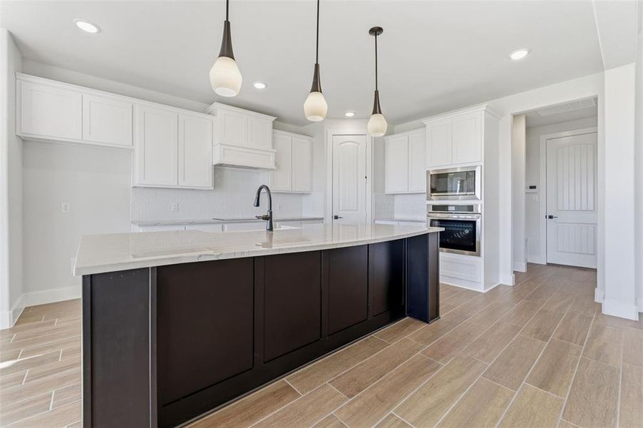 Kitchen with white cabinets, appliances with stainless steel finishes, wood tiled floors, hanging light fixtures, and an island with sink