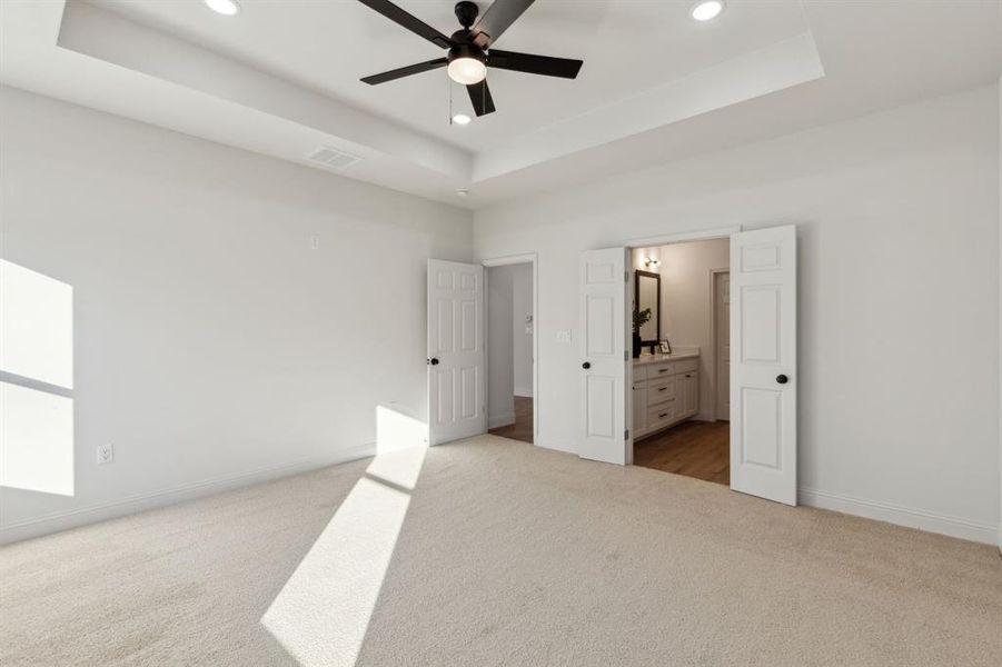 Primary bedroom featuring ensuite bathroom, a raised ceiling, ceiling fan, and light colored carpet