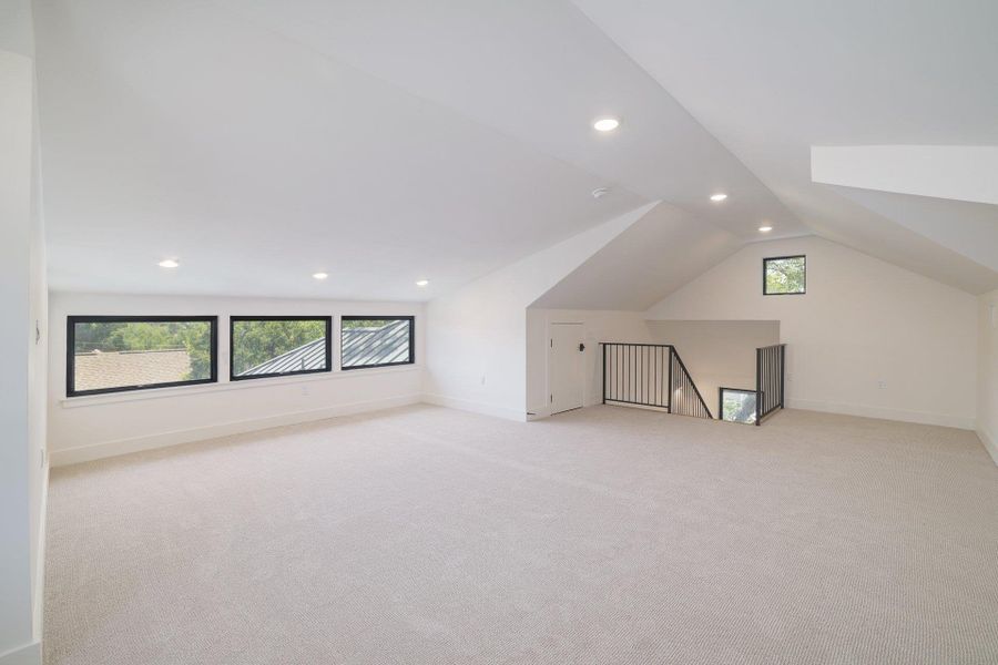 Spacious loft area featuring light-colored carpeting, multiple recessed lights, and a series of three horizontal windows with black frames