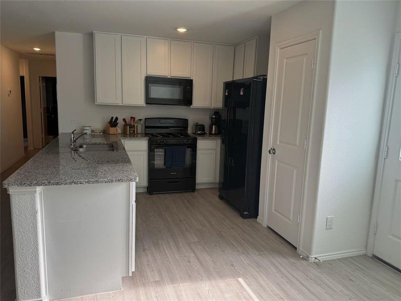 Kitchen featuring an island with a stainless steel sink, granite-style countertop, white cabinetry, black range, and wood-finish flooring Kitchen featuring an island with a stainless steel sink, granite-style countertop, white cabinetry, black range, and wood-finish flooring