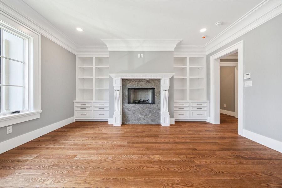 Another view of the primary bedroom room with hardwood floors, a fireplace, built-in shelves, and large windows. Clean, modern design with crown molding and neutral tones.