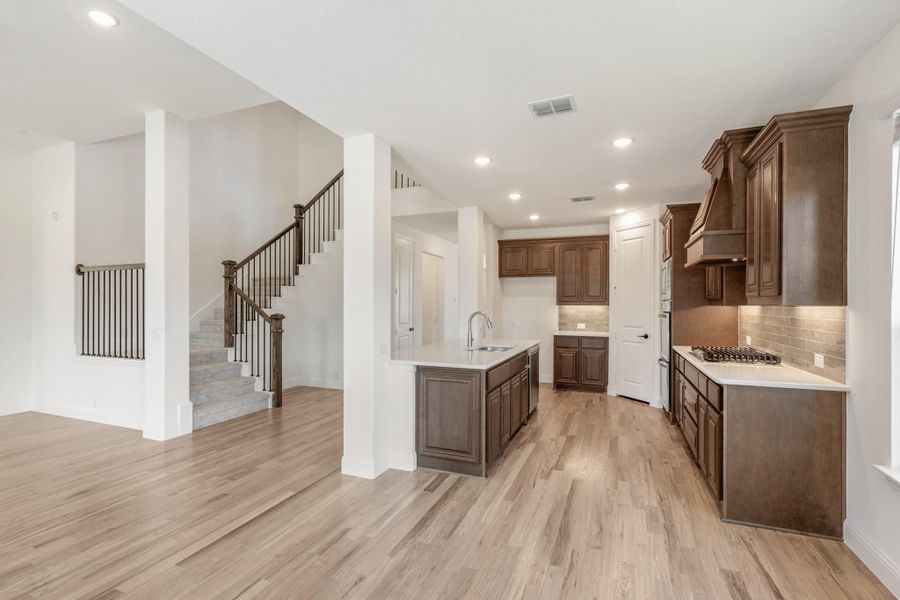 Open kitchen with dark wood cabinets, center island, and light hardwood floors, with staircase visible in background.