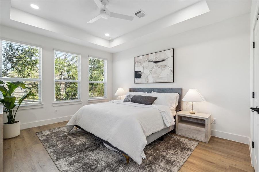 Serene primary bedroom featuring a tray ceiling, large windows for natural light, and a soothing neutral color palette with modern decor for a peaceful retreat.