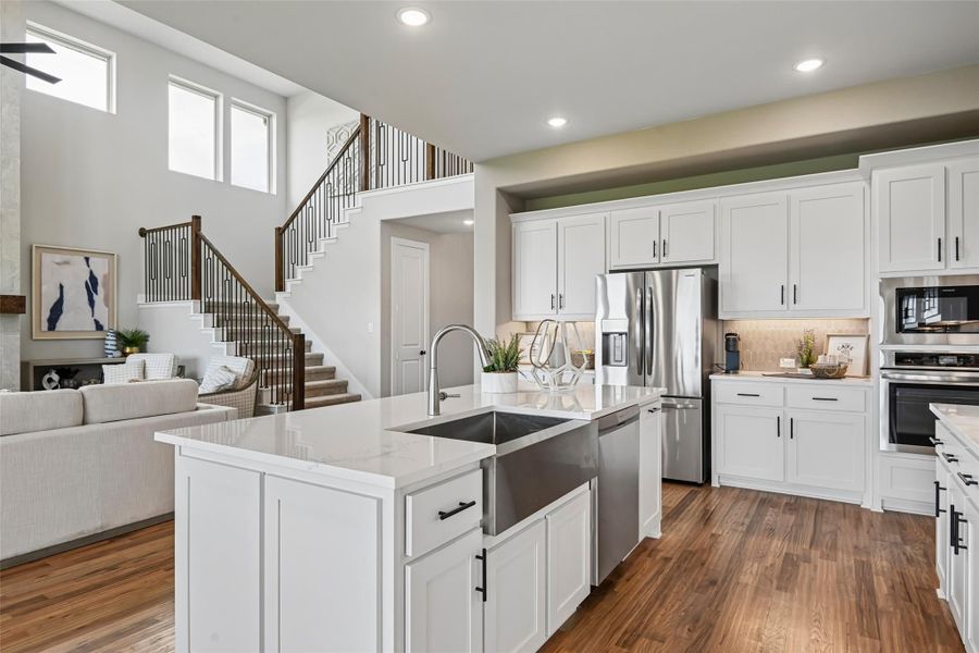 Open-concept kitchen with white cabinets, large island with farmhouse sink, stainless appliances, and hardwood floors overlooking living area and staircase. Open-concept kitchen with white cabinets, large island with farmhouse sink, stainless appliances, and hardwood floors overlooking living area and staircase.