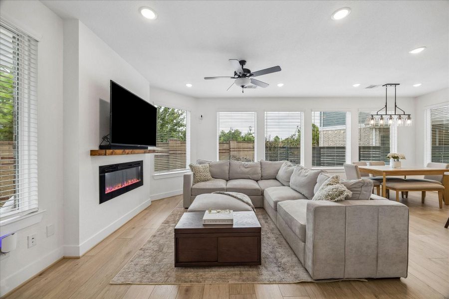 An inviting open-concept living space filled with natural light from a wall of windows, complemented by beautiful Shaw plank flooring throughout. The living room features a sleek electric fireplace, creating a warm and welcoming focal point.