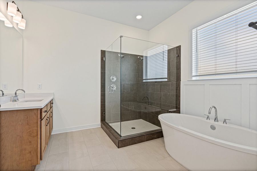 Bathroom featuring a freestanding soaking tub, glass-enclosed shower with dark tile surround, light wood-finish vanity with dual sinks, and board and batten wainscoting Bathroom featuring a freestanding soaking tub, glass-enclosed shower with dark tile surround, light wood-finish vanity with dual sinks, and board and batten wainscoting