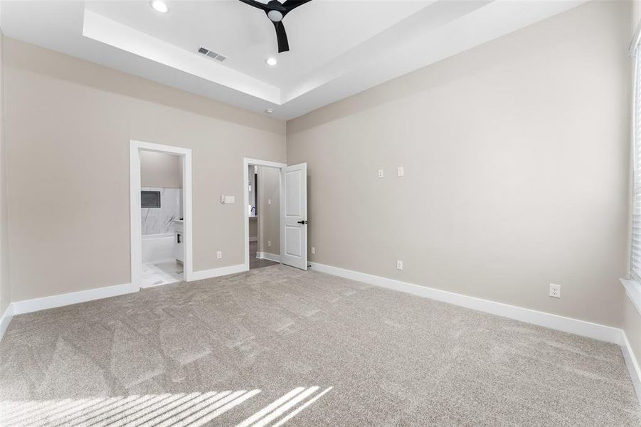 Unfurnished bedroom featuring ceiling fan, ensuite bathroom, a tray ceiling, and light carpet