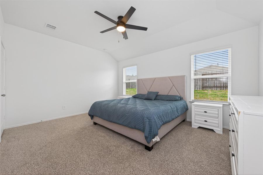 Bright and spacious bedroom with neutral carpeting and walls, featuring a modern ceiling fan and two large windows for natural light.