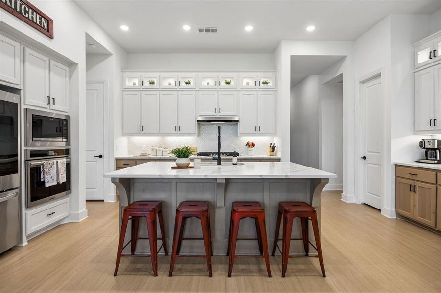 Spacious kitchen featuring a large central island with a stone countertop, light wood-finish flooring, white upper and lower cabinetry, stainless steel appliances, and recessed lighting Spacious kitchen featuring a large central island with a stone countertop, light wood-finish flooring, white upper and lower cabinetry, stainless steel appliances, and recessed lighting