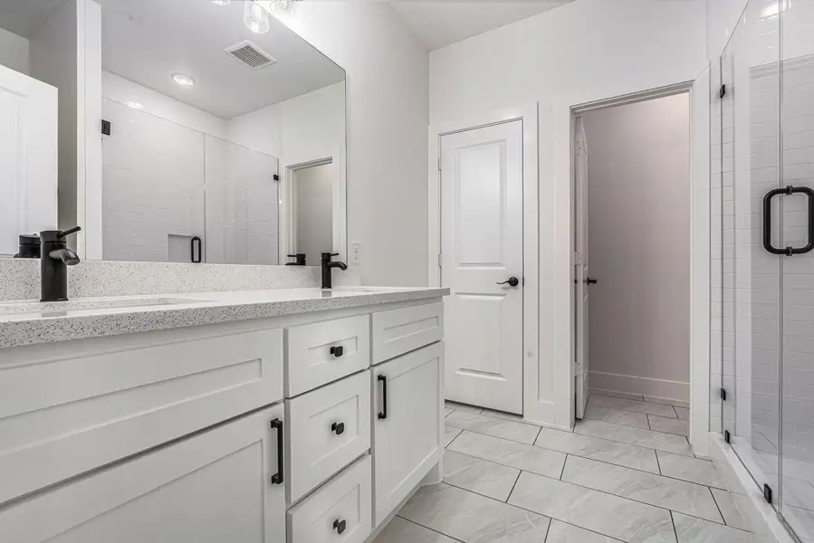This modern bathroom features a spacious double vanity with sleek black fixtures, a large mirror, and a glass-enclosed shower. The neutral color palette and tiled flooring add to the contemporary feel.