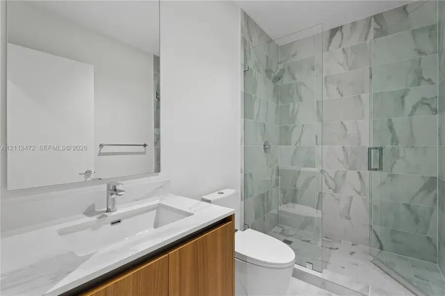 Bathroom featuring a wood-finish vanity with a stone-top integrated sink, a large wall-mounted mirror, a walk-in shower with floor-to-ceiling tile, and a glass partition