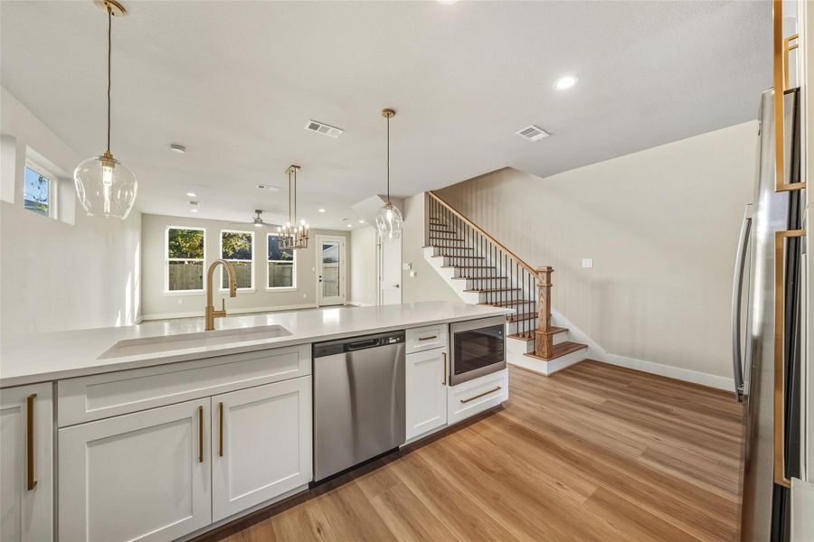 This modern kitchen features a spacious open layout with sleek white cabinetry, stainless steel appliances, and a large island with pendant lighting. It seamlessly flows into a bright living area with wood flooring and large windows, complemented by a staircase leading to the upper level.