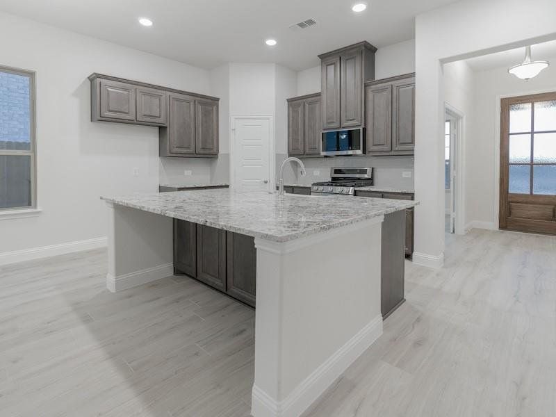 Kitchen with tasteful backsplash, appliances with stainless steel finishes, a kitchen island with sink, light stone countertops, and light hardwood / wood-style floors