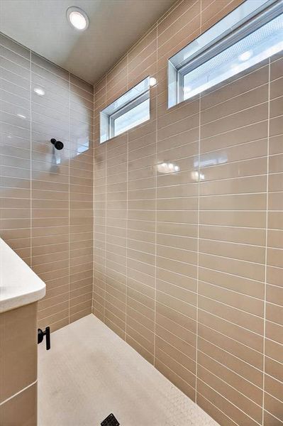 Walk-in shower featuring rectangular beige wall tiles, small hexagonal floor tiles, black shower fixtures, two high horizontal windows, and recessed ceiling lighting Walk-in shower featuring rectangular beige wall tiles, small hexagonal floor tiles, black shower fixtures, two high horizontal windows, and recessed ceiling lighting