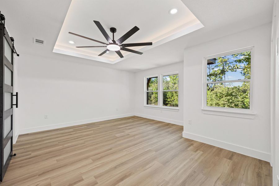 Primary bedroom with a tray ceiling, light wood-type flooring, recessed lighting, a barn door, and a ceiling fan