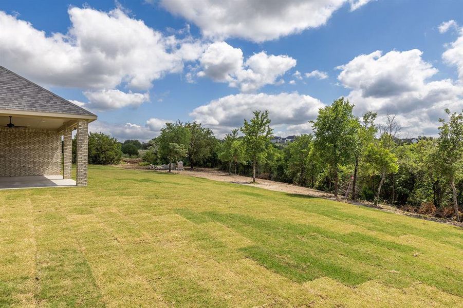 Expansive lawn with a covered patio featuring a ceiling fan, brick exterior, and mature trees bordering the property