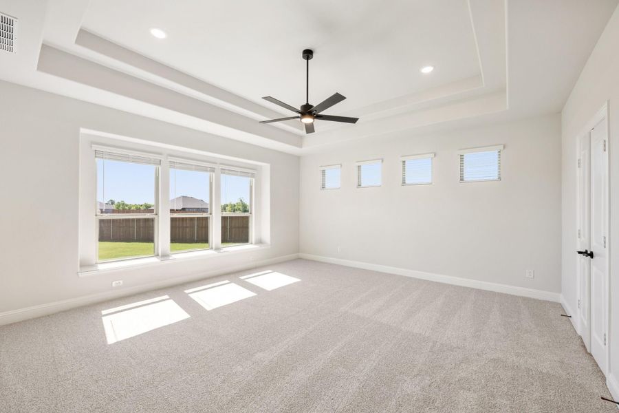 Empty bedroom with tray ceiling, ceiling fan, carpet flooring, and large windows overlooking a backyard Empty bedroom with tray ceiling, ceiling fan, carpet flooring, and large windows overlooking a backyard