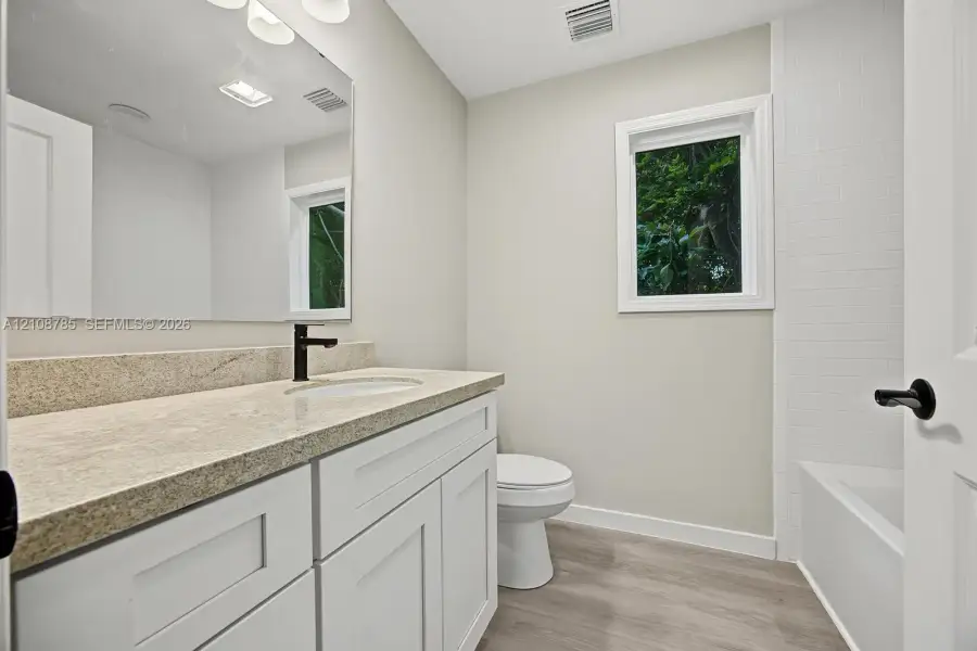 Bathroom featuring a white shaker-style vanity, stone-surface countertop, dark metal hardware, wood-finish flooring, and a subway-tile tub surround Bathroom featuring a white shaker-style vanity, stone-surface countertop, dark metal hardware, wood-finish flooring, and a subway-tile tub surround