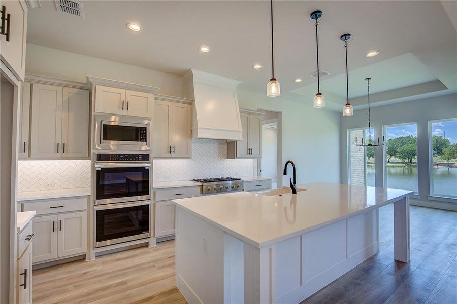 Kitchen with custom range hood, light hardwood / wood-style flooring, backsplash, a raised ceiling, and appliances with stainless steel finishes