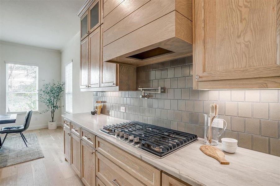 Kitchen featuring light hardwood flooring, a kitchen island with leathered Quartzite countertops, farm sink, decorative backsplash, premium range hood, stainless steel gas stovetop, pot-filler,  and natural stain custom cabinetry with soft close drawers and cabinets.