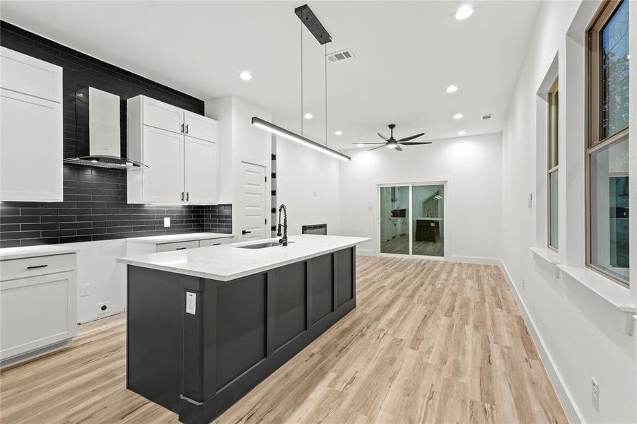 Kitchen with ceiling fan, hanging light fixtures, wall chimney range hood, a kitchen island with sink, and white cabinets