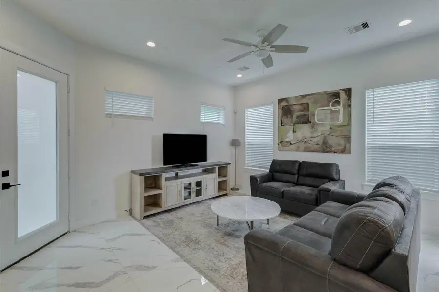Bright furnished living room with soaring ceilings, recessed lighting, ceiling fan, marble-look tile flooring, large windows with blinds, and comfortable seating. A stylish media console, area rug, and contemporary wall art create an inviting space for relaxing or entertaining.