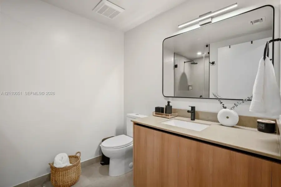 Wood-finish vanity cabinet with a stone-surface countertop, rectangular undermount sink, matte black faucet, and large rectangular mirror with rounded edges