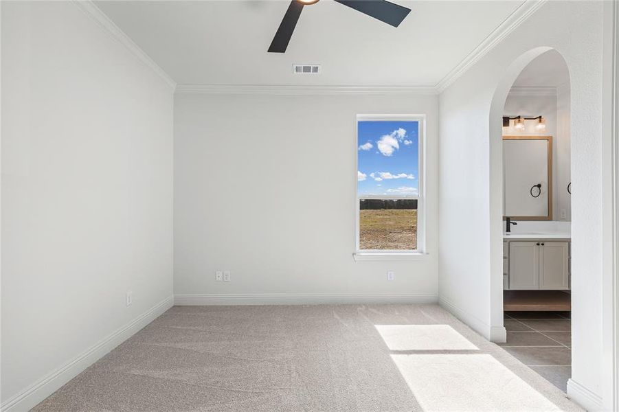 Unfurnished bedroom with arched walkways, light carpet, ceiling fan, ensuite bath, and ornamental molding