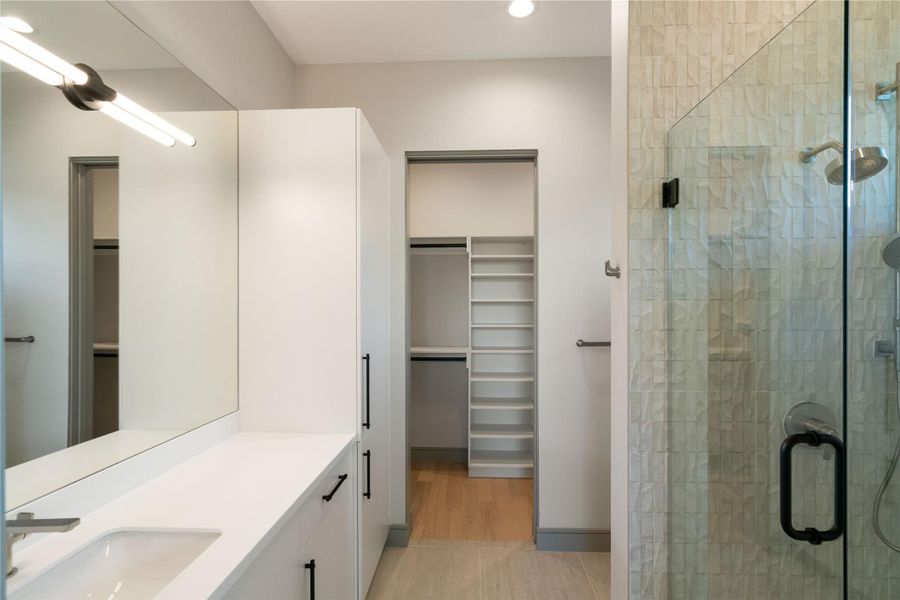 This modern bathroom features a sleek design with a large mirror, a white vanity with ample storage, a glass-enclosed shower, and an adjoining walk-in closet with built-in shelving.