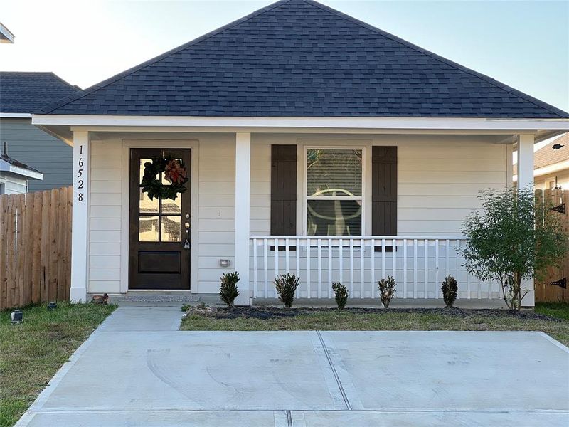 This is a single-story home featuring a gabled roof, a covered front porch with railing, a dark front door, and a fresh concrete driveway. The facade combines white siding with dark window shutters, complemented by a neatly landscaped front yard.