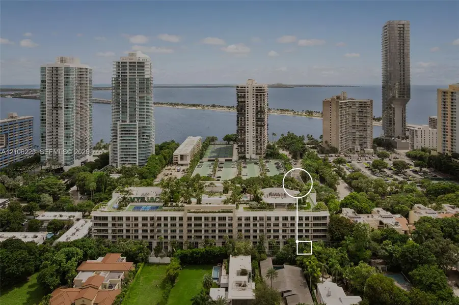 Aerial perspective of a multi-story building complex featuring a rooftop tennis court, mature tree canopy, proximity to the coastline, and surrounding high-rise structures Aerial perspective of a multi-story building complex featuring a rooftop tennis court, mature tree canopy, proximity to the coastline, and surrounding high-rise structures