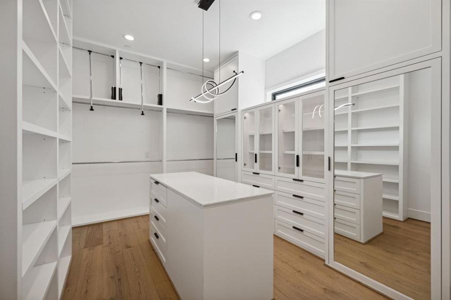 The closet features ample storage space with floor-to-ceiling shelving and hanging rods on both sides. A large central island with drawers provides additional storage.The closet is finished with a white oak wood floor and a marble countertop on the island.