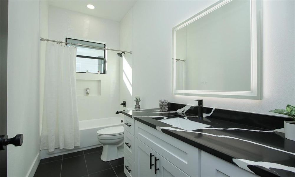 This bathroom features a modern design quartz countertop, white cabinetry, and a large illuminated mirror. It includes a bathtub with a shower combination, white tiles, and a black-framed window for natural light.