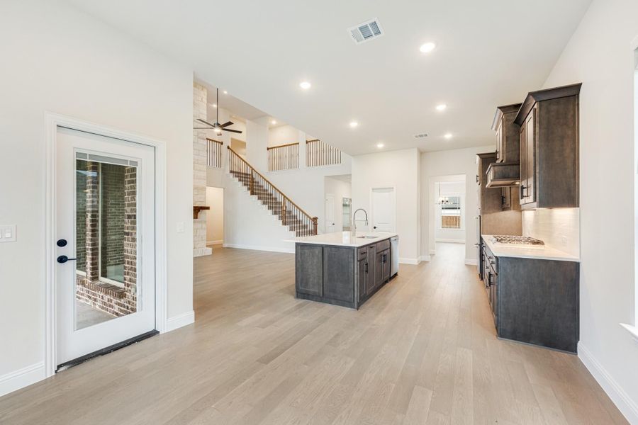 Open-concept kitchen with dark cabinets, center island, and light wood floors, open to living area with staircase and high ceilings Open-concept kitchen with dark cabinets, center island, and light wood floors, open to living area with staircase and high ceilings