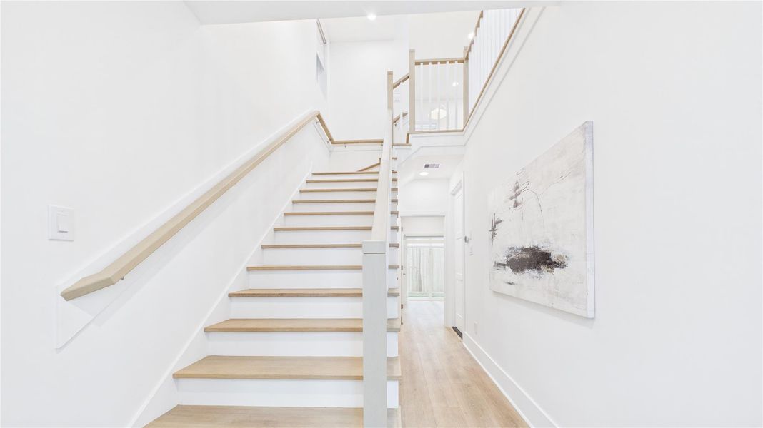 This photo showcases a bright, modern staircase with light wood steps and sleek white walls. An abstract painting adds a touch of elegance to the hallway, which features natural light from a window at the end.