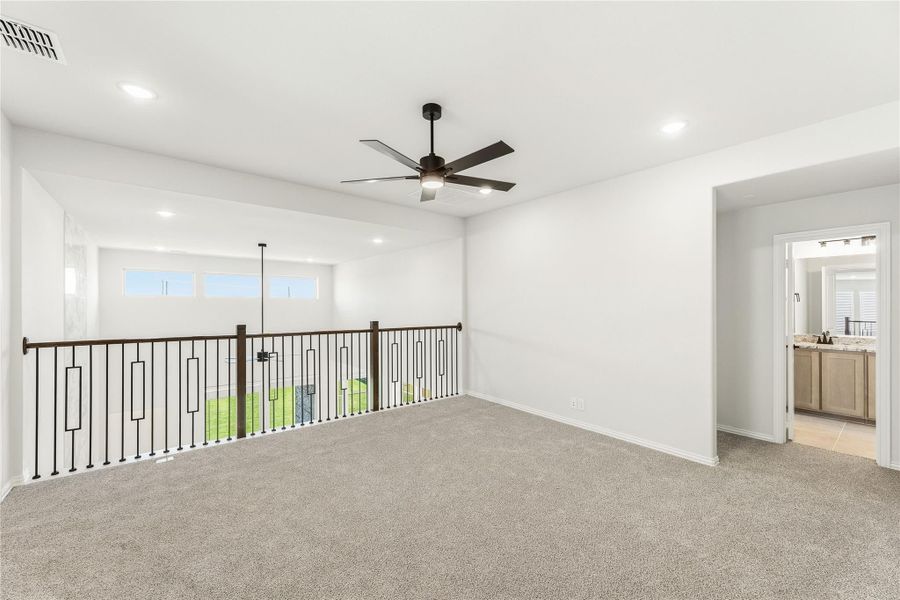 Open upstairs loft with carpet, ceiling fan, metal railing overlooking lower level, and adjacent bathroom doorway.