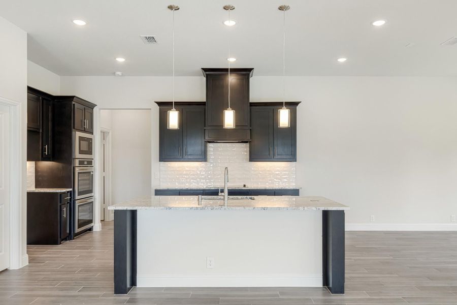 Kitchen with dark cabinets, marble island, white tile backsplash, and three pendant lights above the sink Kitchen with dark cabinets, marble island, white tile backsplash, and three pendant lights above the sink