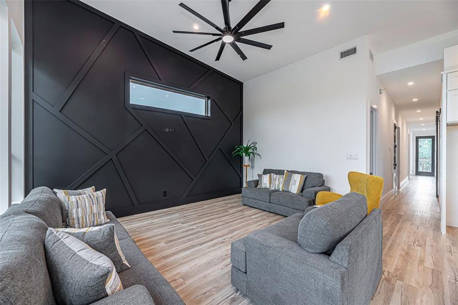 The home showcases a modern living room with a striking black accent wall featuring geometric patterns, complemented by a contemporary ceiling fan. Light hardwood floors extend throughout, leading to a bright hallway.