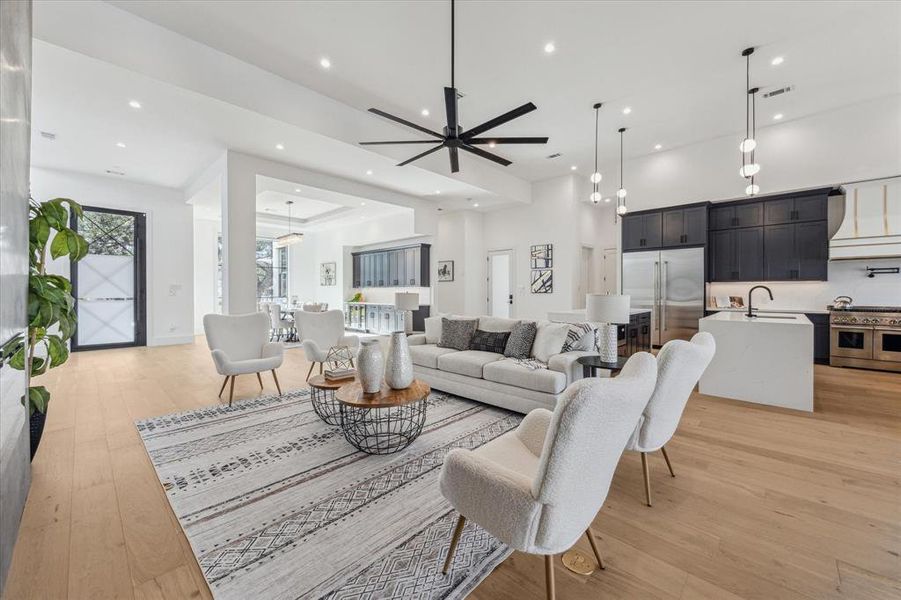 This family room features an open-concept design with high ceilings, recessed lighting, light wood flooring, and a modern black ceiling fan. It seamlessly connects to the kitchen and dining areas, creating a spacious and inviting layout.