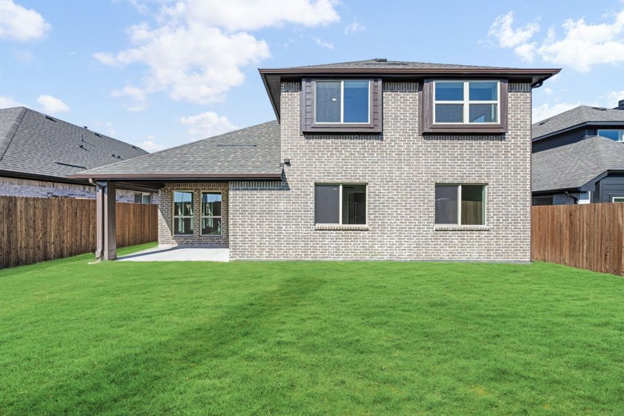 Two-story brick home rear exterior with covered patio, large green lawn, and wood privacy fence