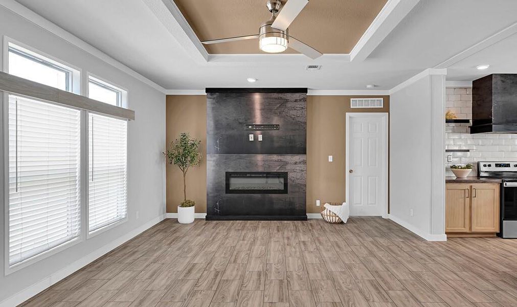 Living area featuring a ceiling fan, a raised ceiling, light wood-type flooring, a fireplace, and crown molding Living area featuring a ceiling fan, a raised ceiling, light wood-type flooring, a fireplace, and crown molding