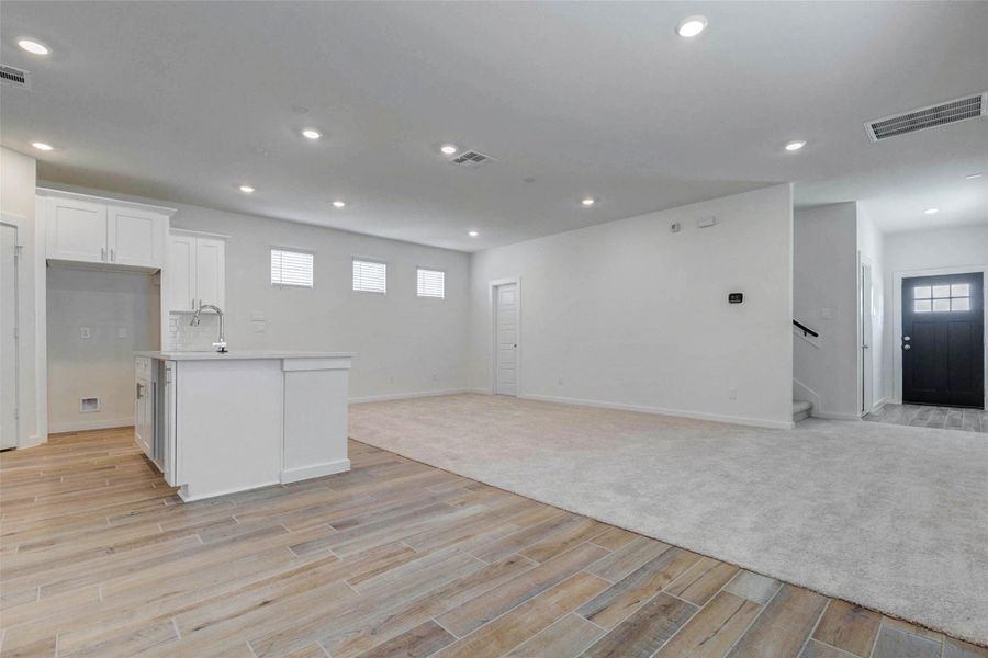Wide open view from the kitchen into the living area, showing the island, white cabinetry, wood-look tile, carpeted living space, recessed lighting, and front entry beyond. The layout provides easy flow for entertaining while keeping the main living spaces connected.