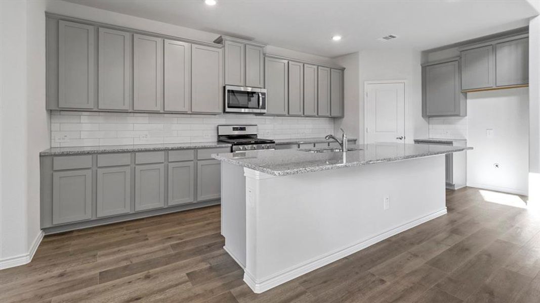 Kitchen with gray cabinets, backsplash, light stone countertops, appliances with stainless steel finishes, and a kitchen island with sink