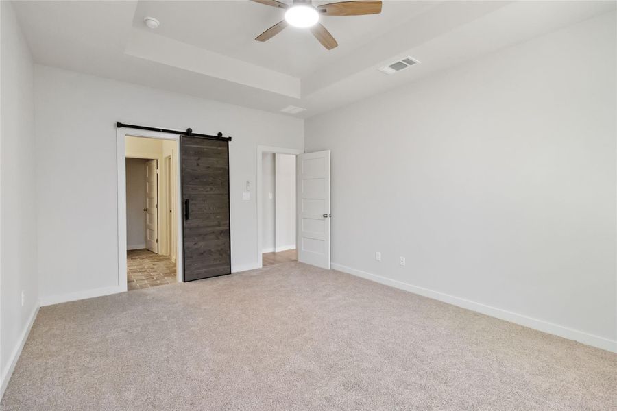 Unfurnished bedroom with light colored carpet, a tray ceiling, a barn door, and a ceiling fan Unfurnished bedroom with light colored carpet, a tray ceiling, a barn door, and a ceiling fan